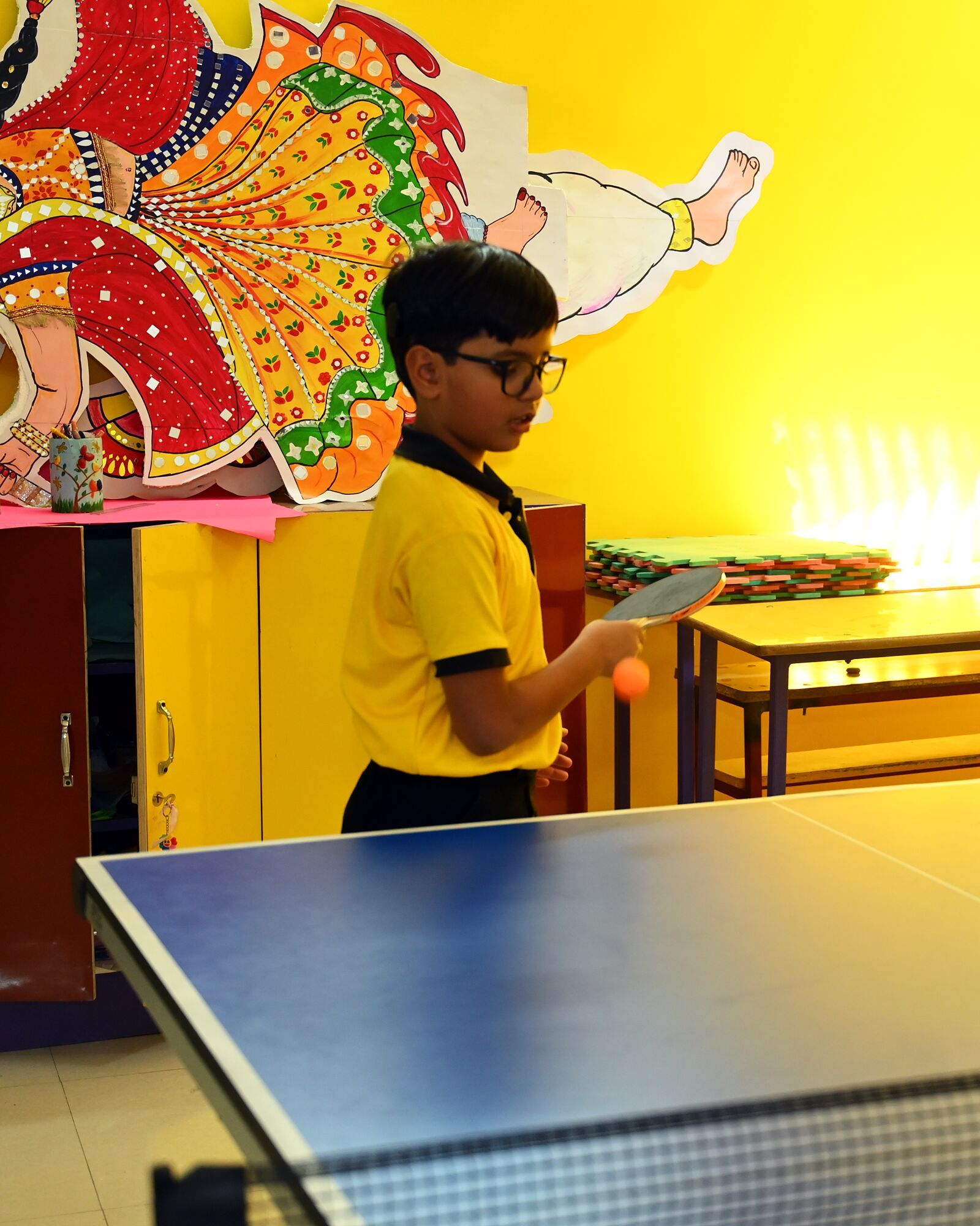 A boy in glasses at the table tennis table, bat in one hand and the ball in the other, ready to serve