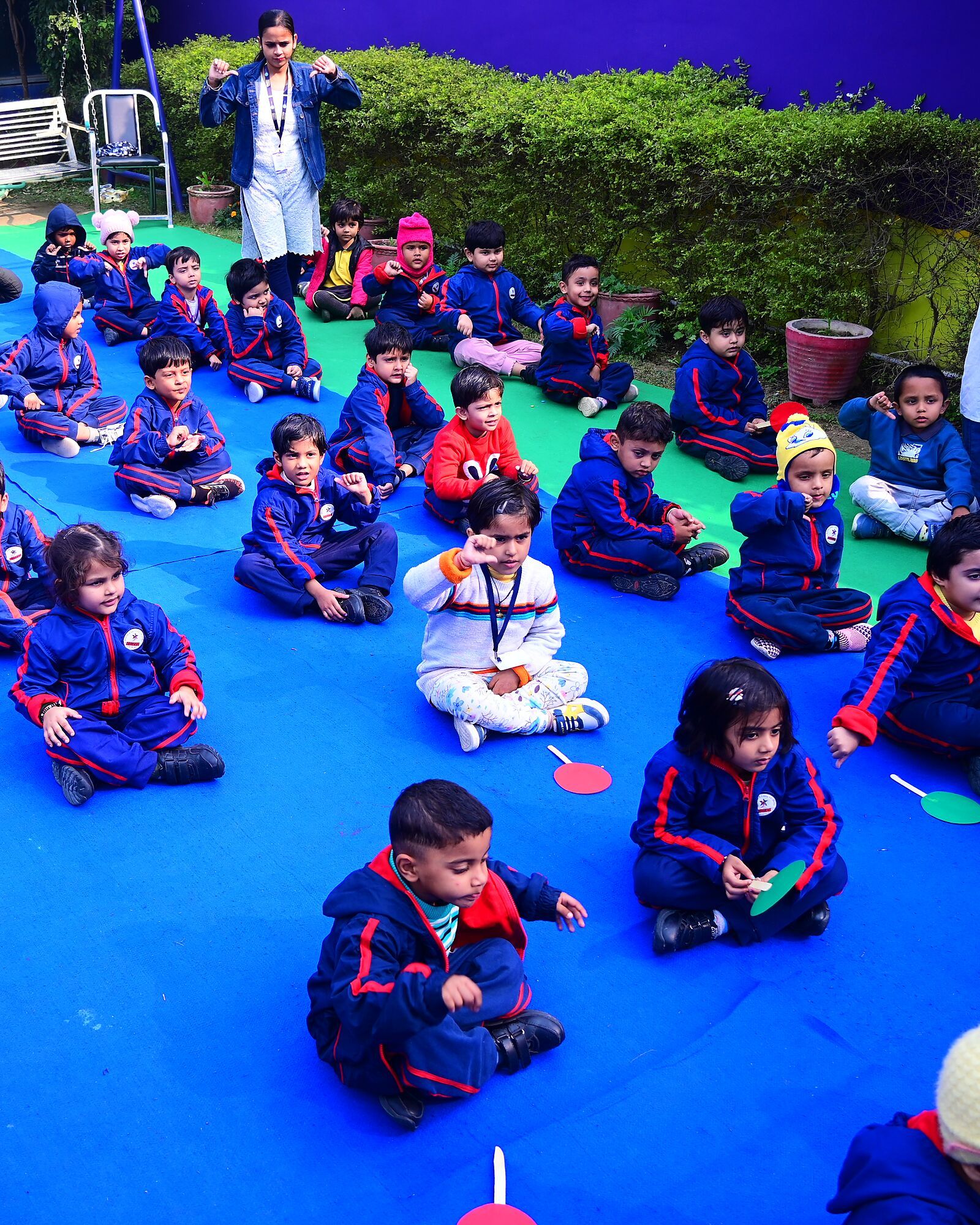 Children sitting cross-legged on mats outdoors for yoga, a teacher standing by