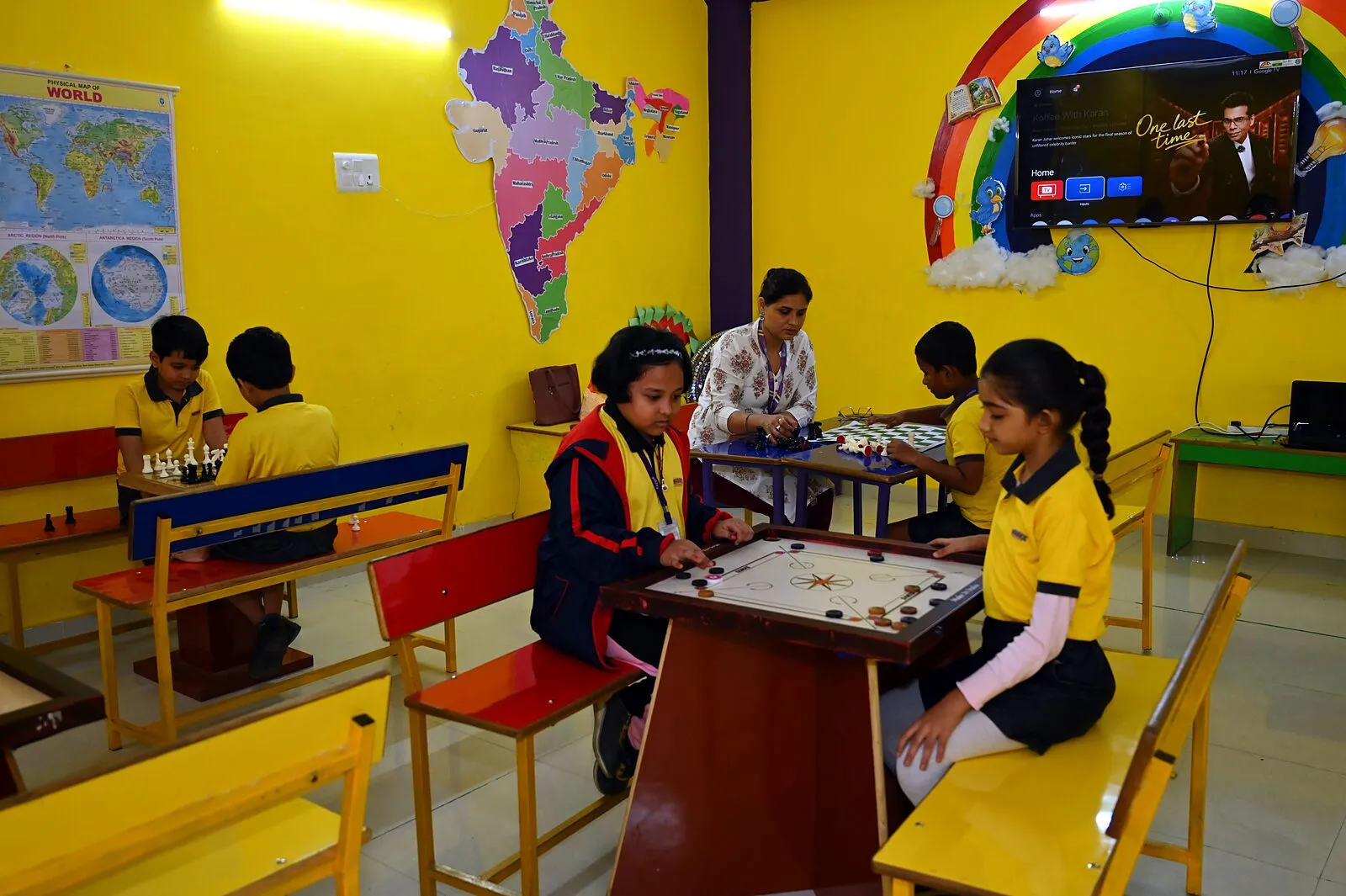 The indoor games hall: children at carrom and at chess, a teacher with them, a map of India on the wall