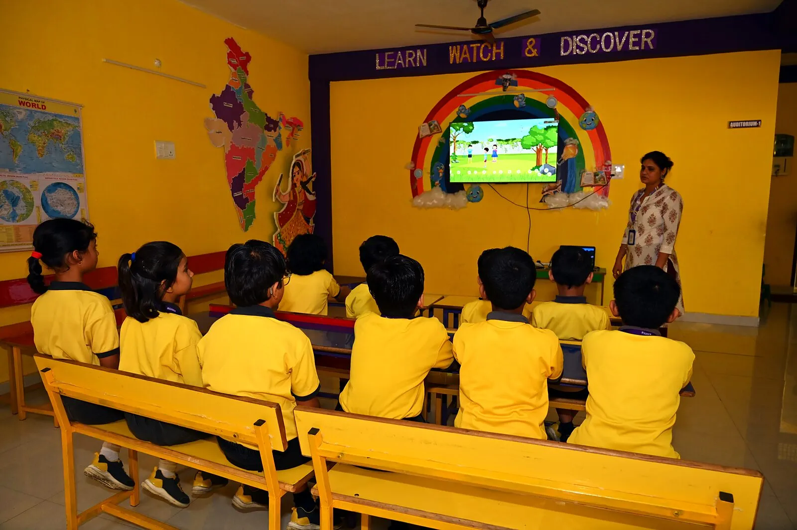 Children on benches from behind, watching a learning programme on the big screen under the words Learn, Watch & Discover