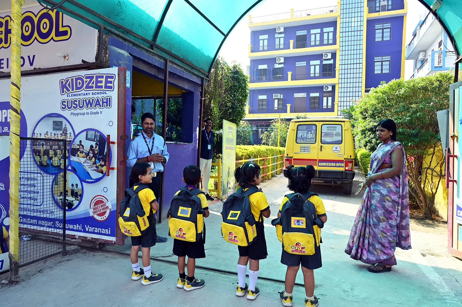 The gate in the morning: the guard on duty, children with their bags walking in, a member of staff beside them