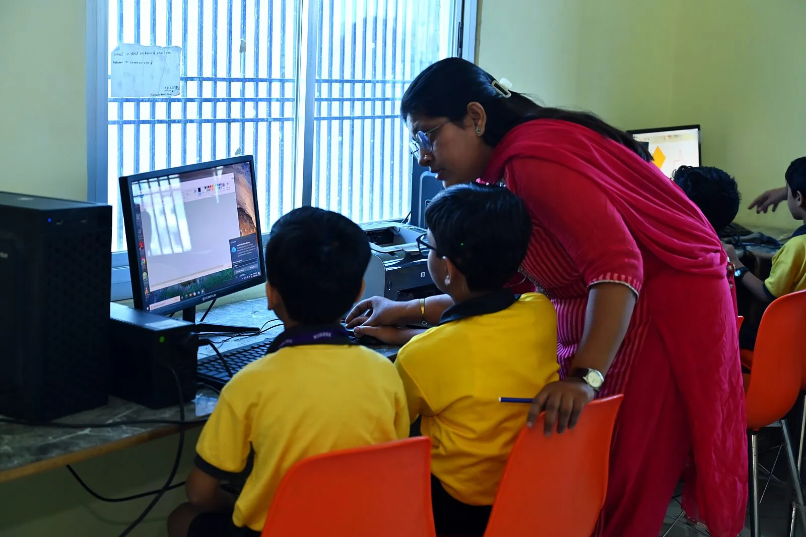 A teacher leaning in beside two boys at a computer, guiding one of them