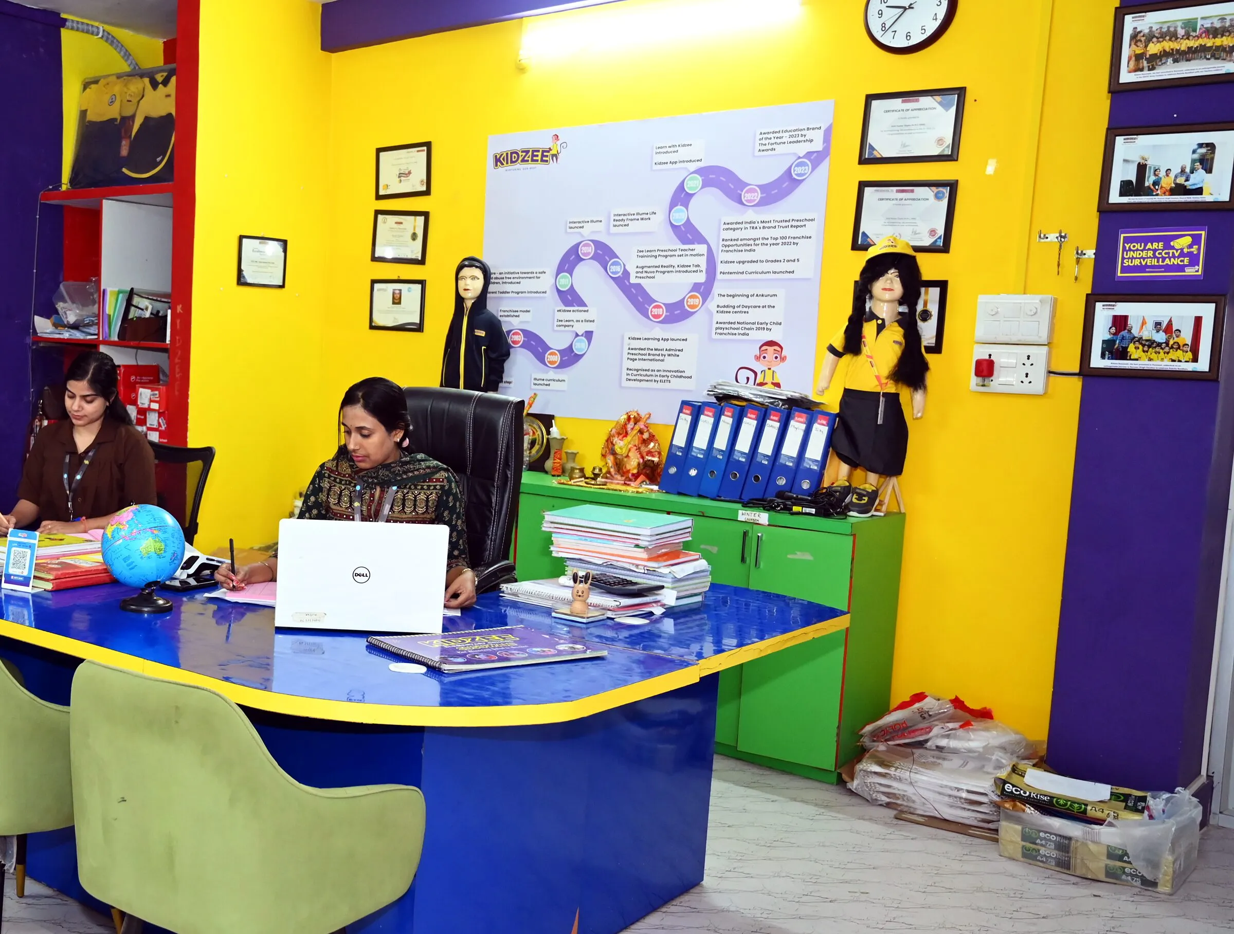 The reception: the long blue desk, two of the office staff at work, the yellow wall of certificates behind