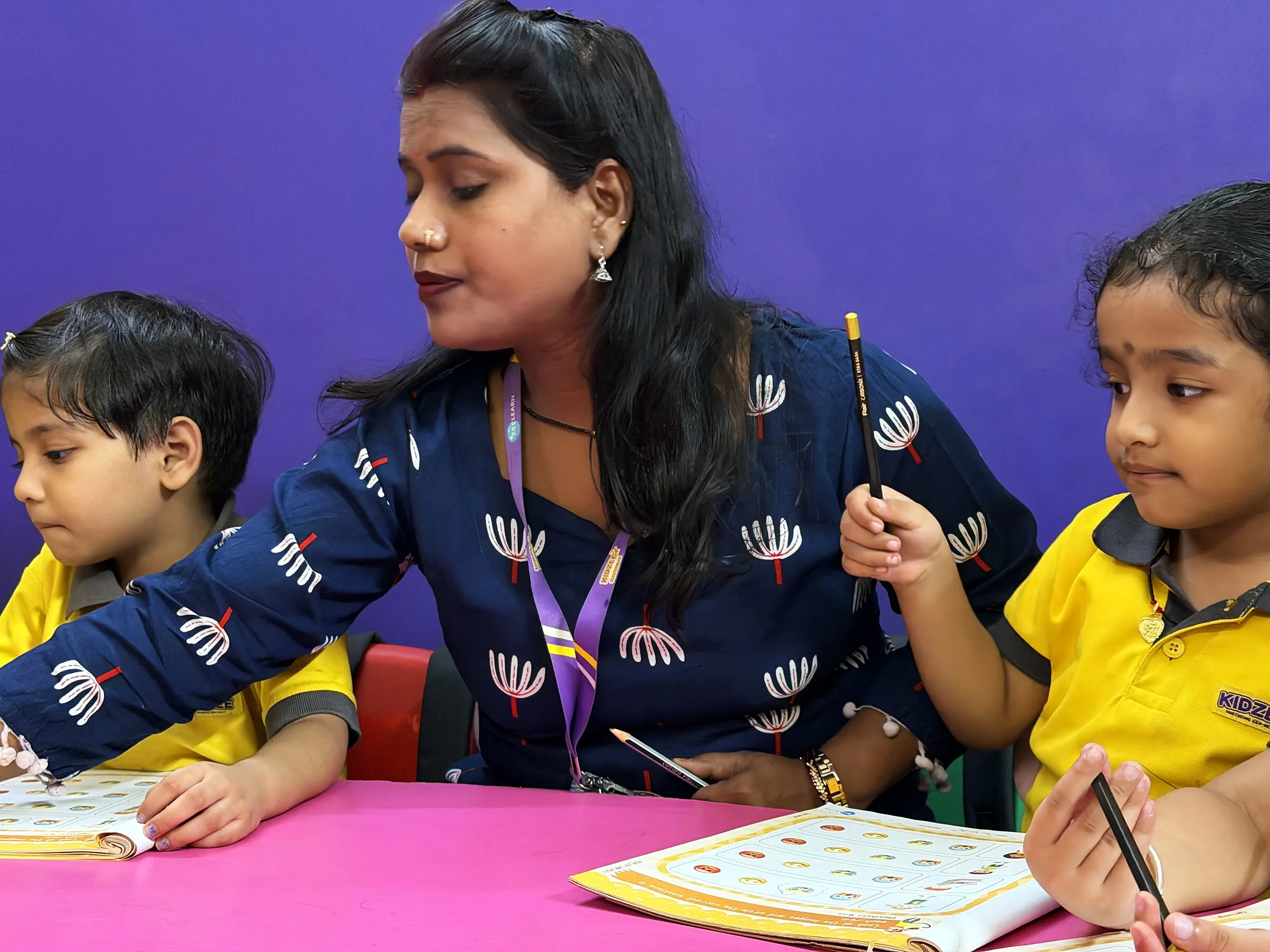 A teacher leaning in to help two children with their worksheets at a pink table