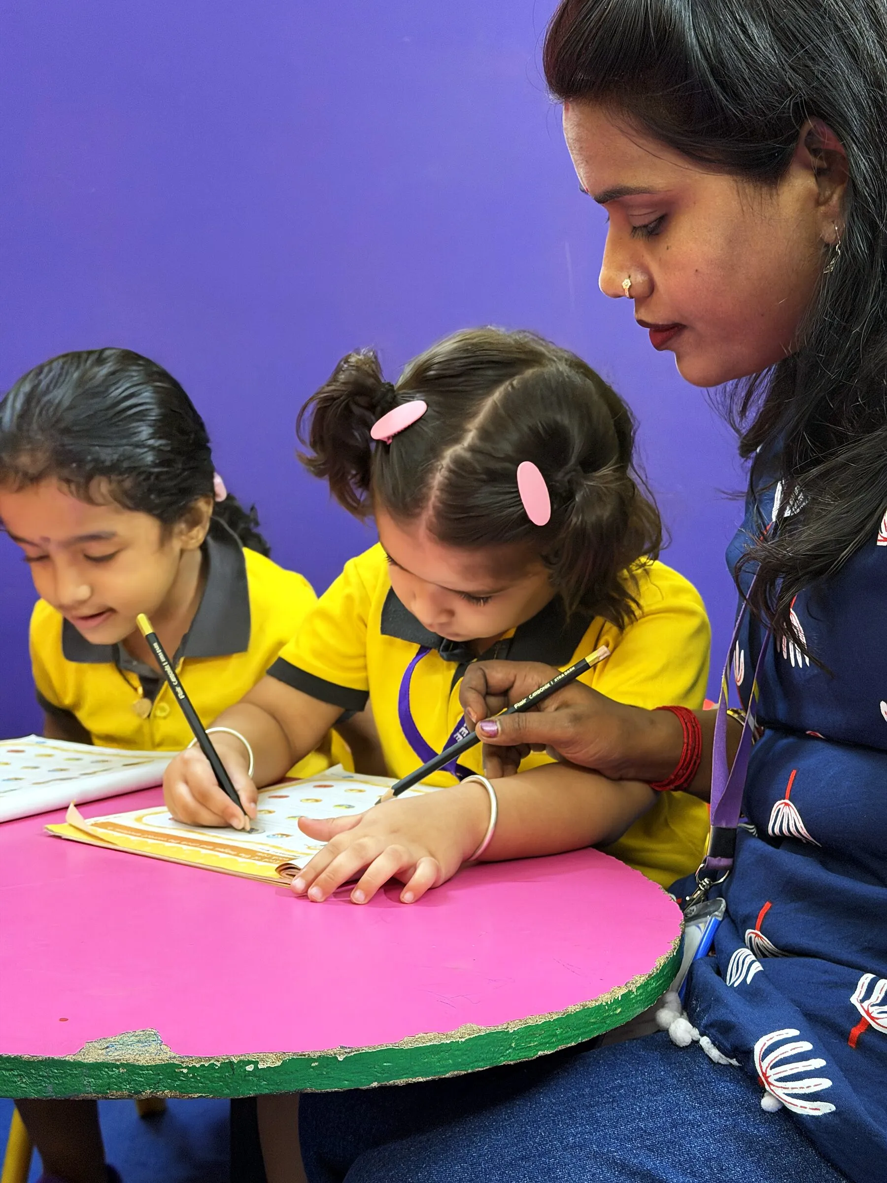 Two girls writing on their worksheets in pencil, their teacher beside them