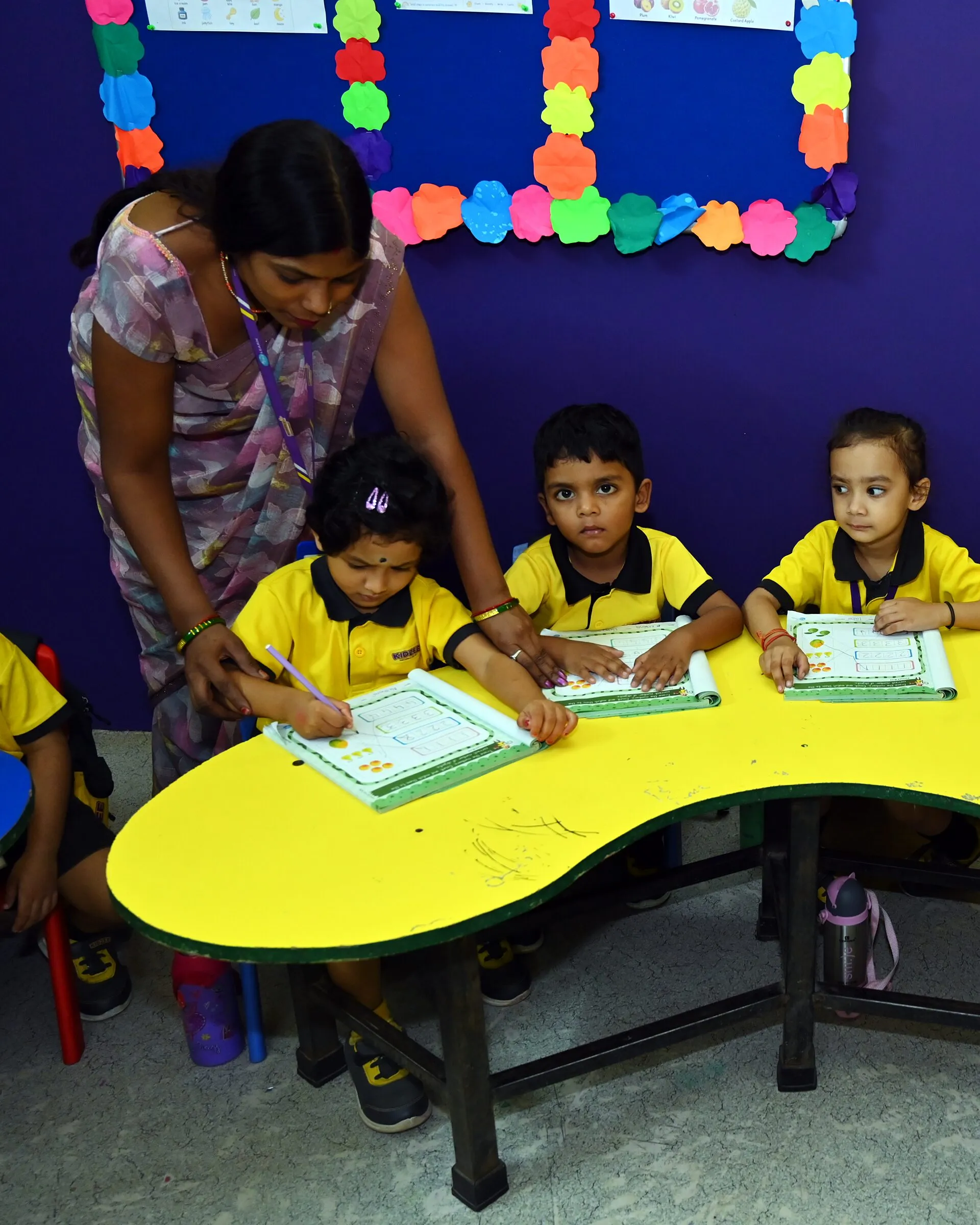 A teacher guiding a girl's hand as she writes in her number book, two classmates beside her at the yellow table