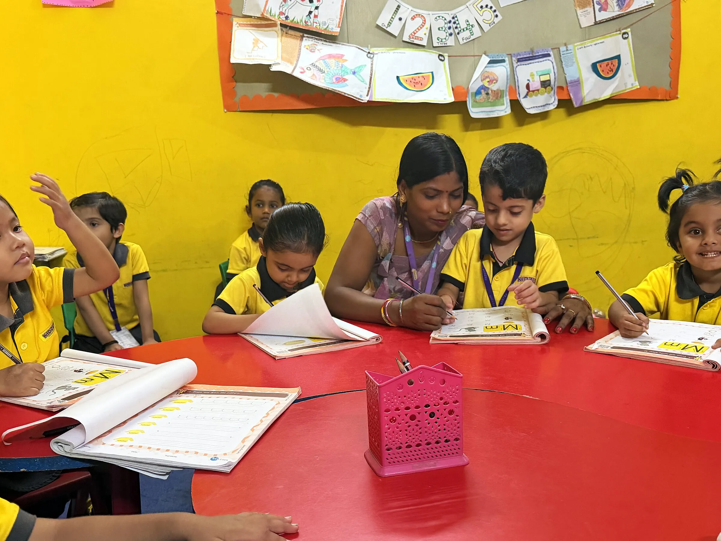 Children writing in their workbooks round a red table, the teacher guiding a boy's pencil