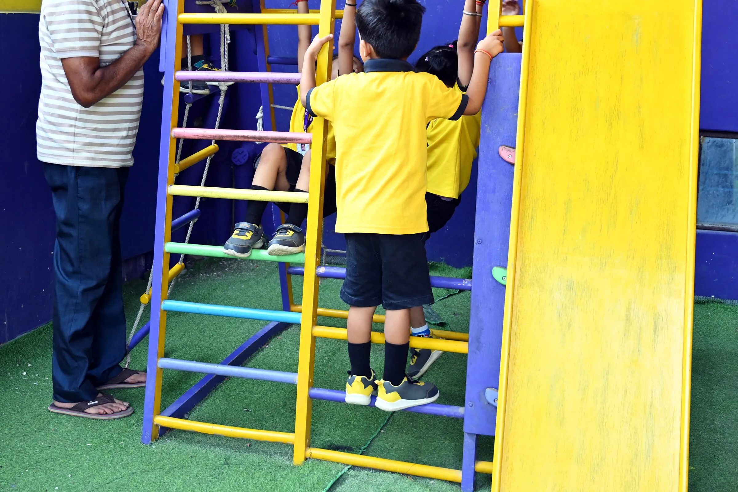 A boy climbing the frame beside the slide, seen from behind, a grown-up standing right beside him