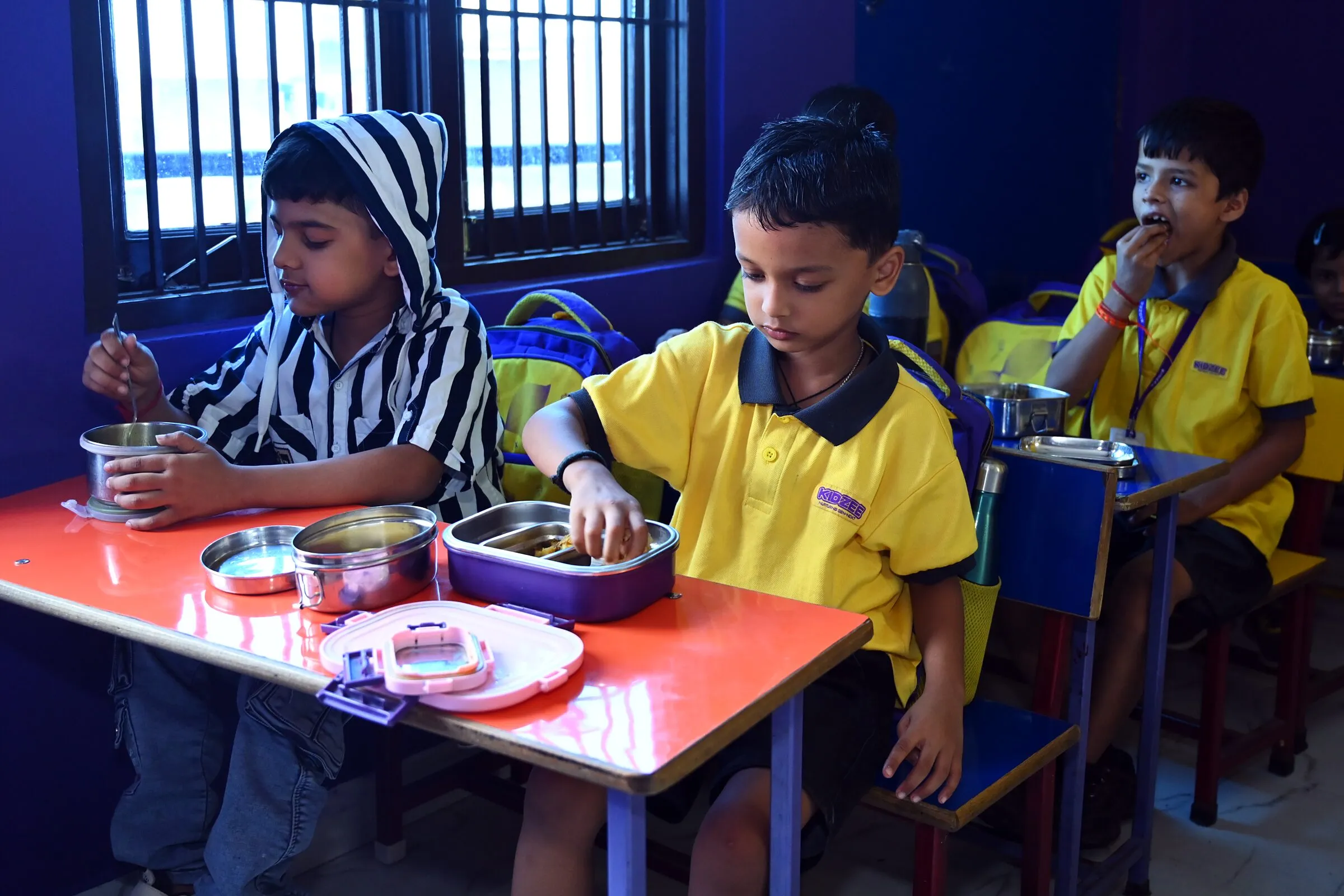 Children at their desks by the window eating from their tiffin boxes