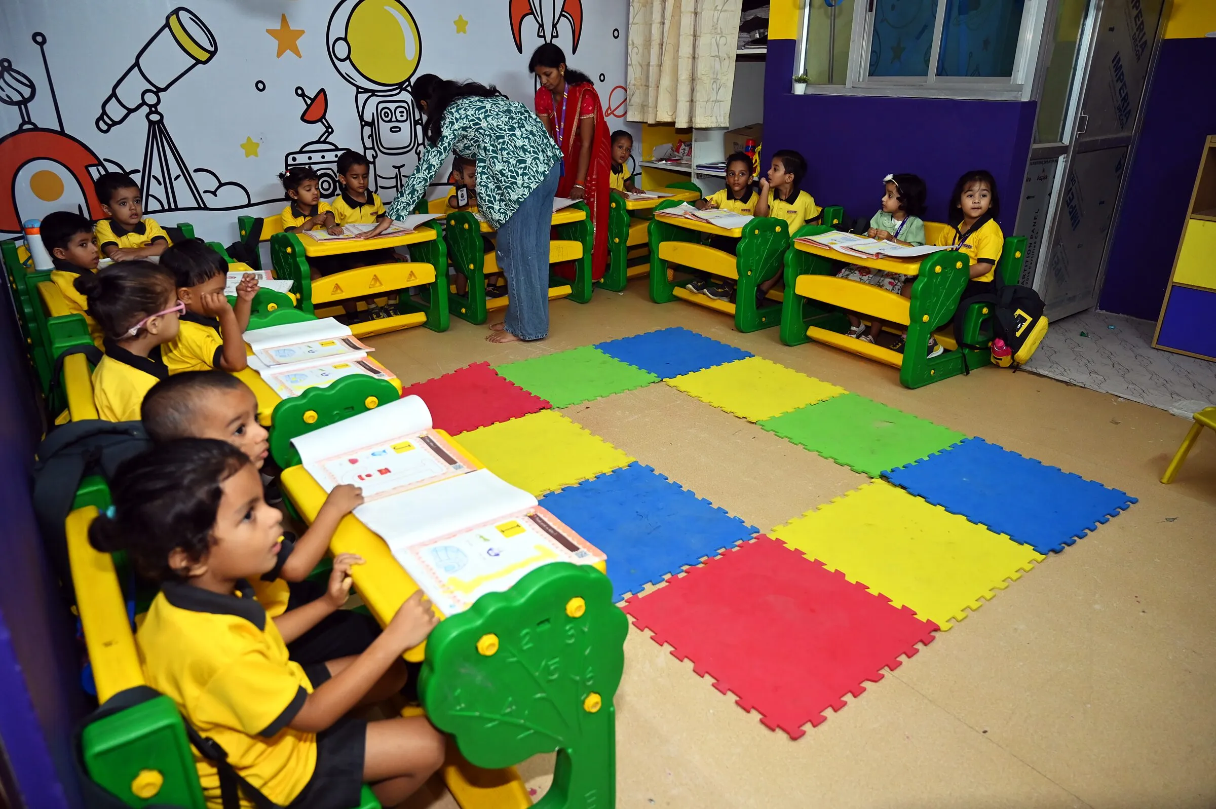 The class settled at their desks round the floor mats, workbooks open, a teacher bending to help at the back