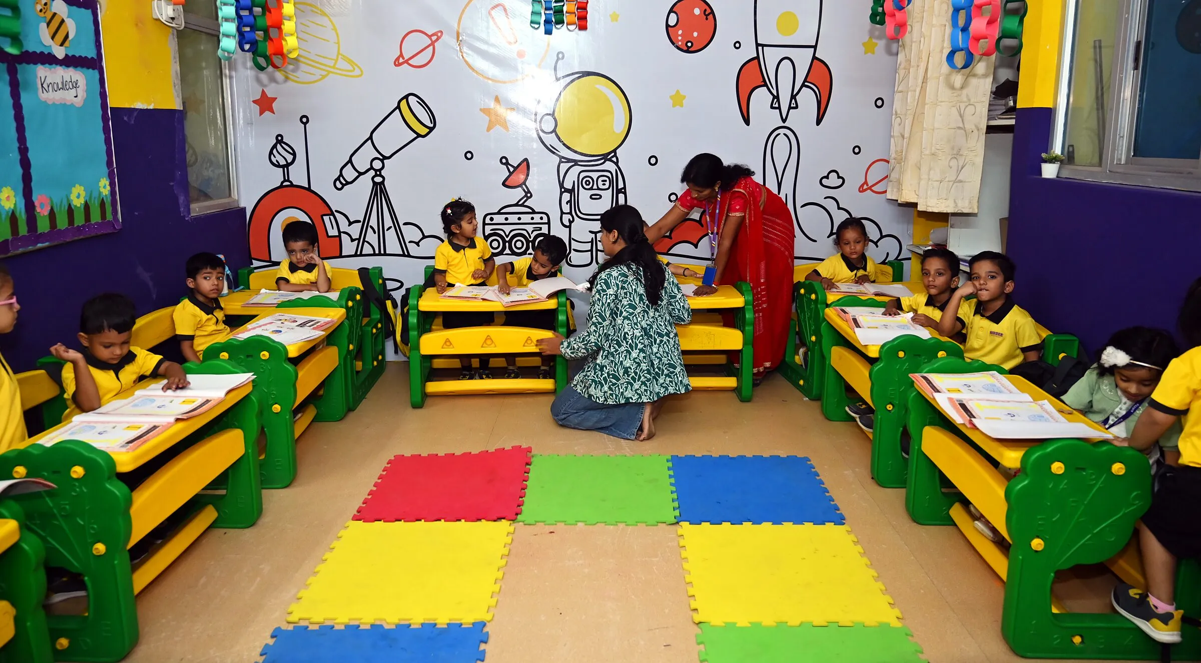 The class at their desks round the floor mats under a space mural, a teacher kneeling with two children
