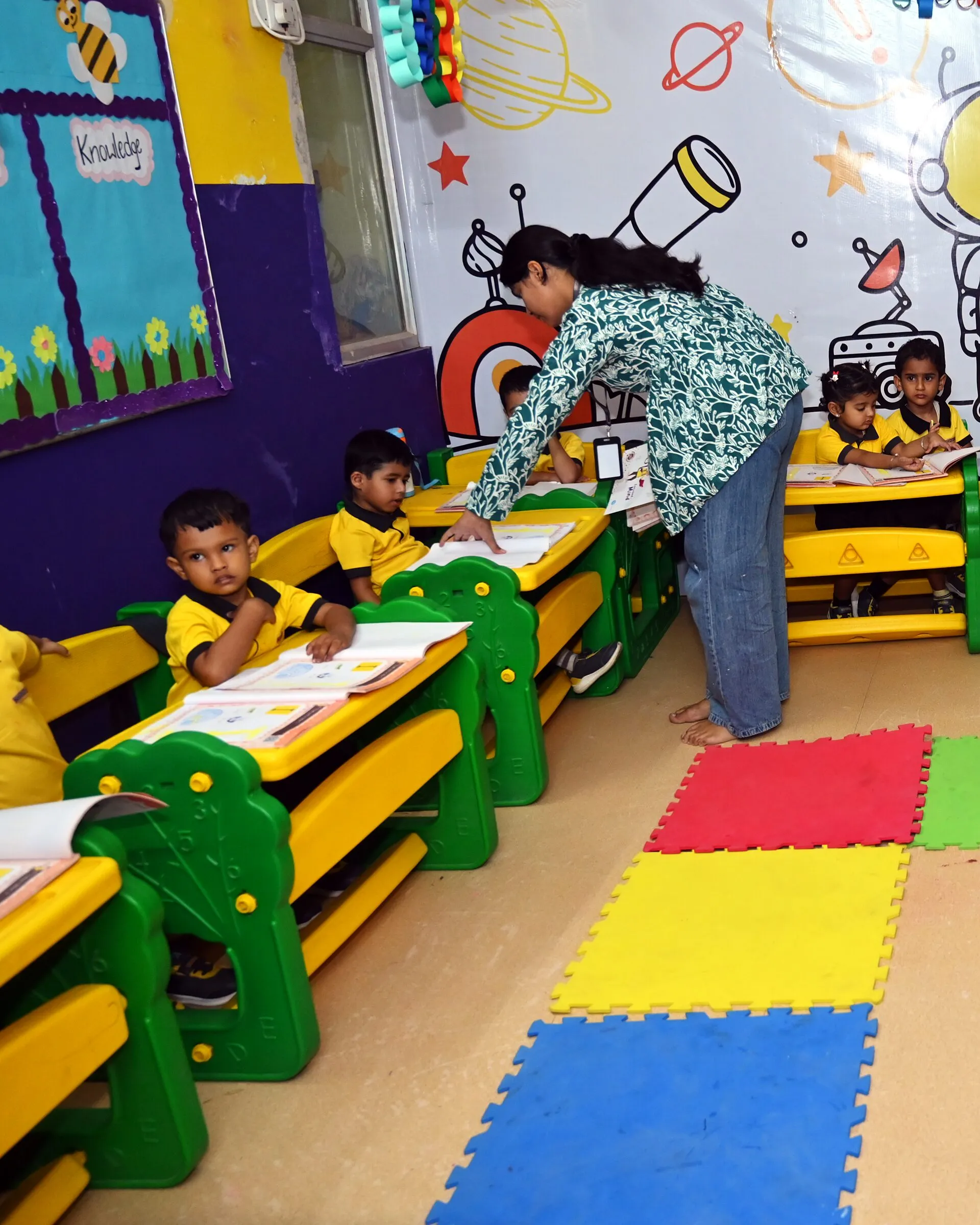 A teacher bending over a boy's open book at his desk, his classmates at their books around him
