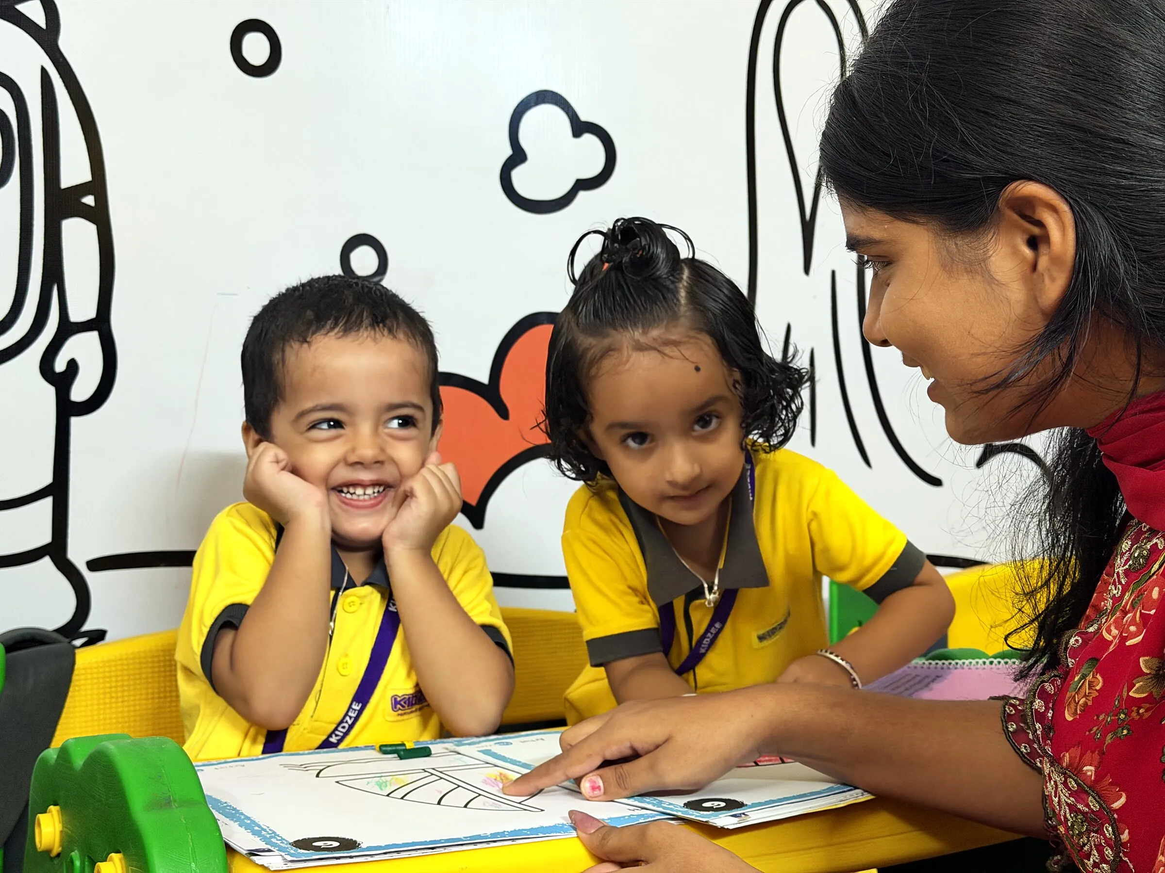 Two little ones at a worksheet with their teacher, one beaming with his chin in his hands
