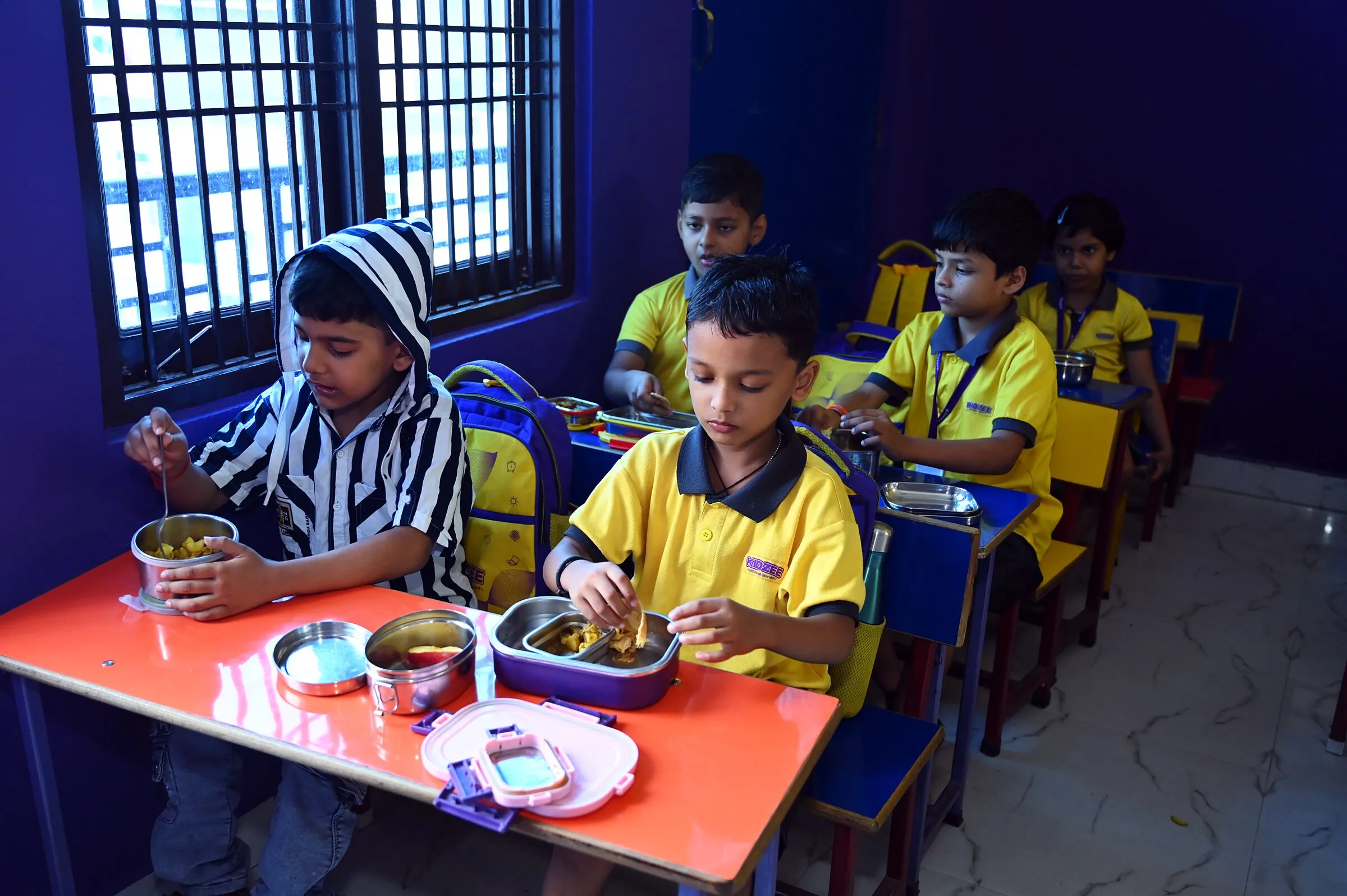 Two boys at an orange table eating from their open tiffin boxes, the class at their desks behind