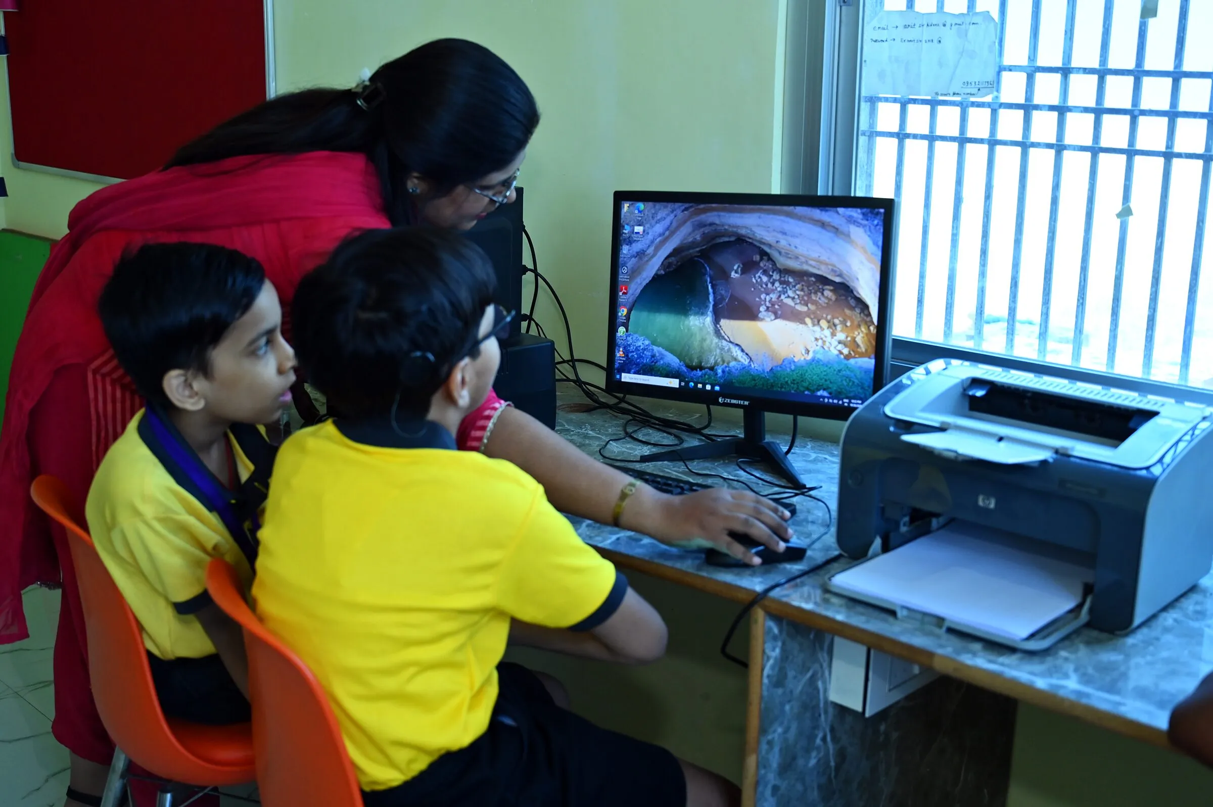 A teacher leaning in to help two boys at a computer, a picture on the screen