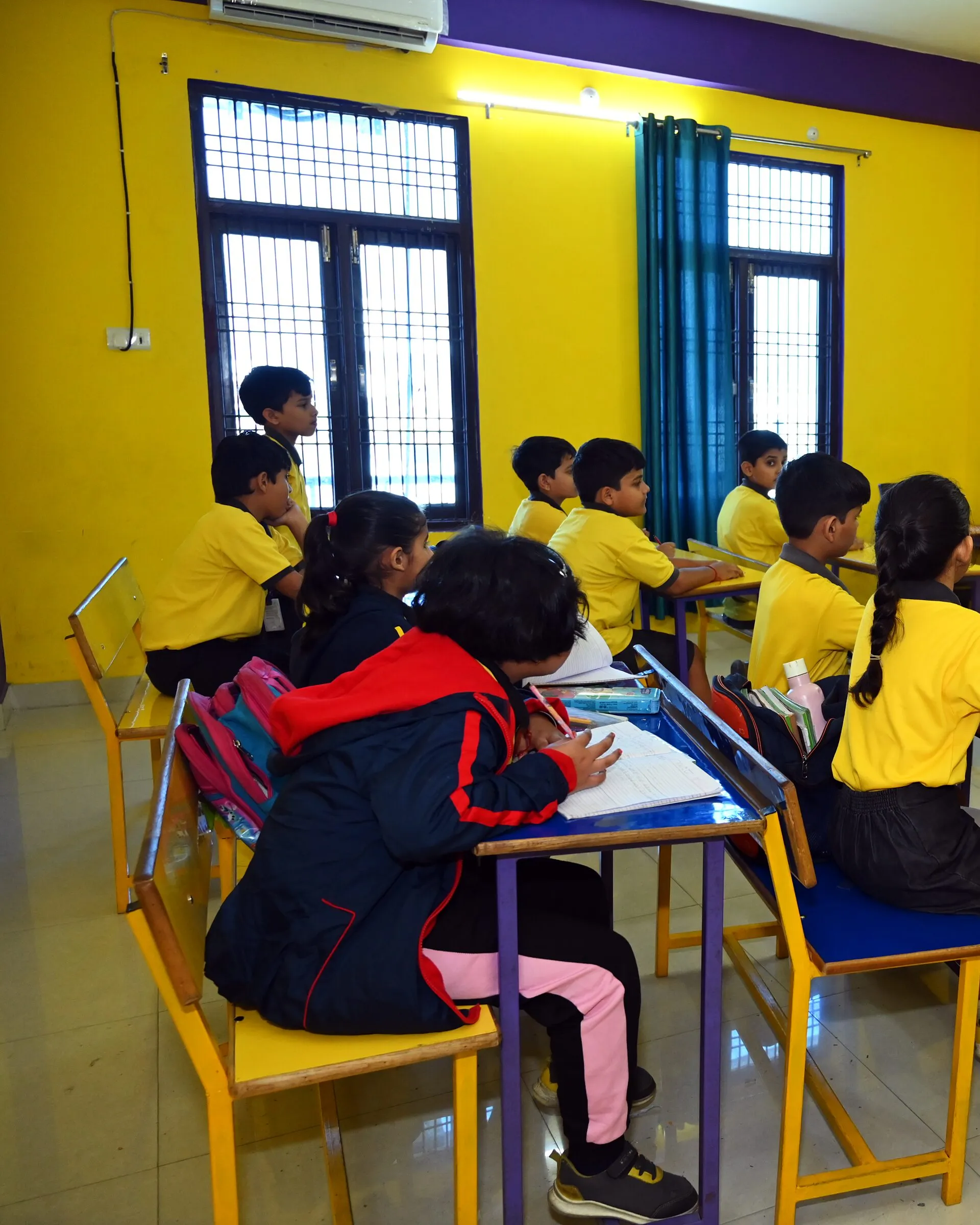 Children writing at their desks by the windows, seen from behind, a girl in a red jacket at the front