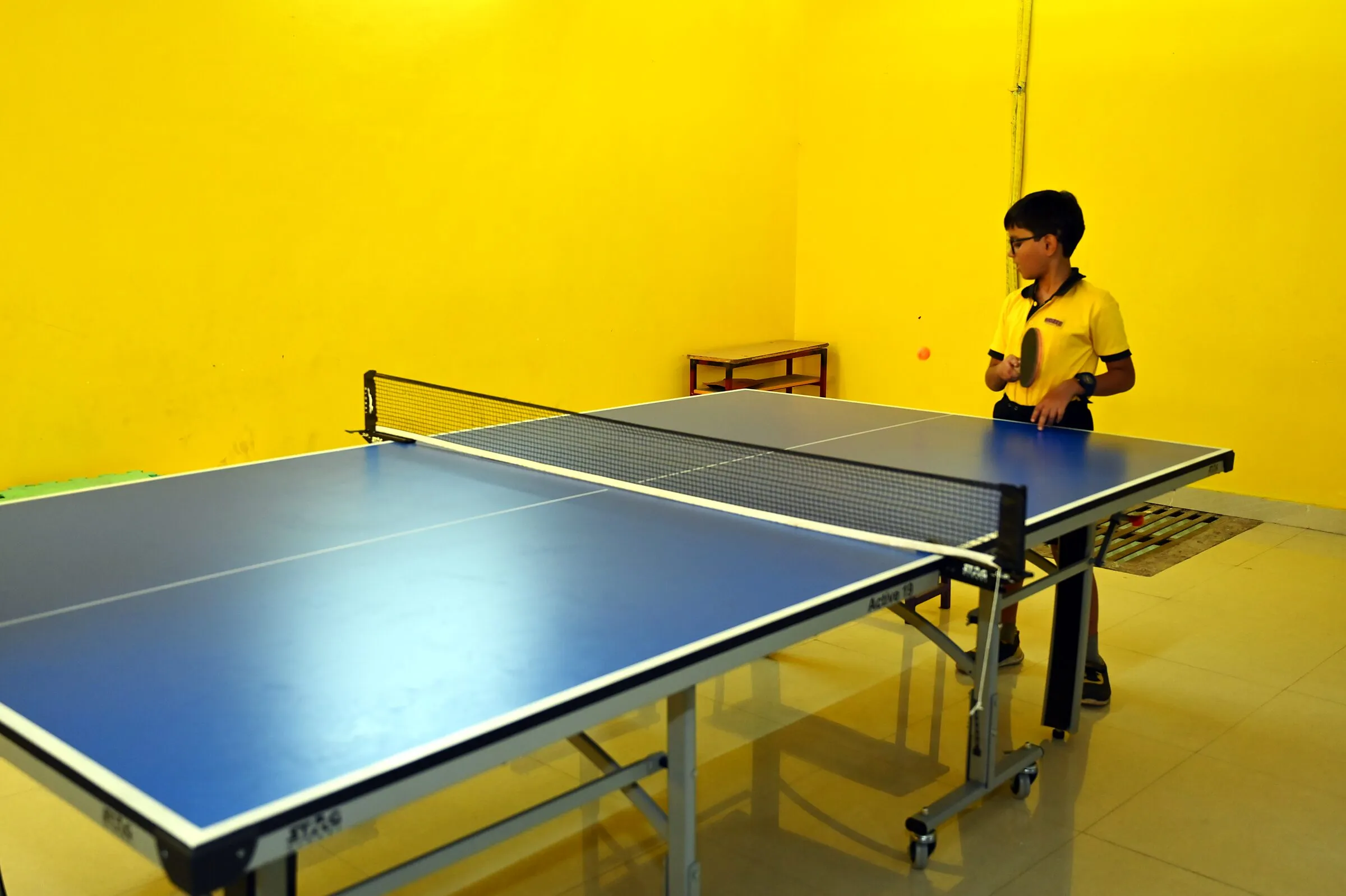 A boy at the table tennis table, bat ready, the room wide and yellow around him