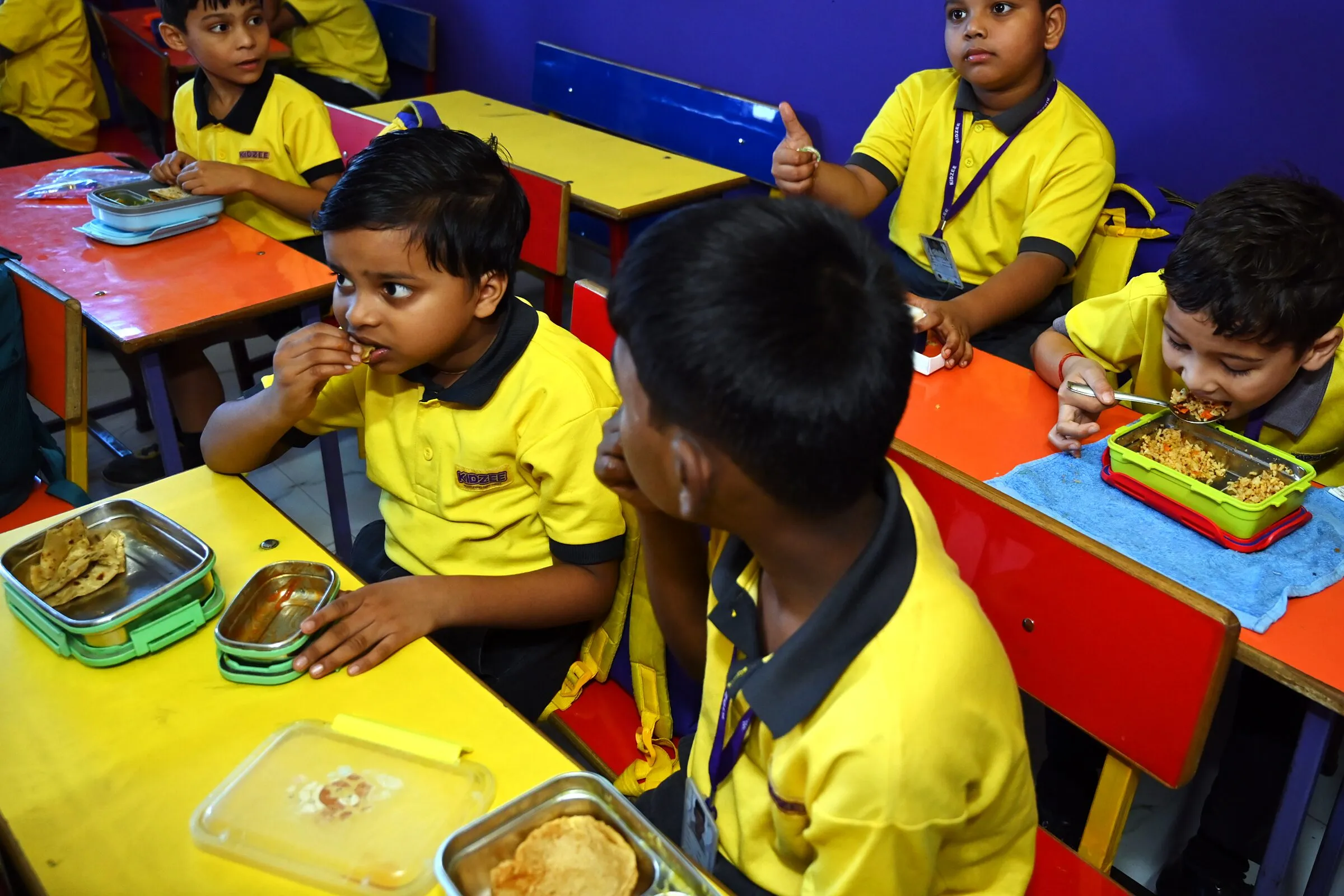 Boys at their desks eating from their tiffin boxes, one giving a thumbs up