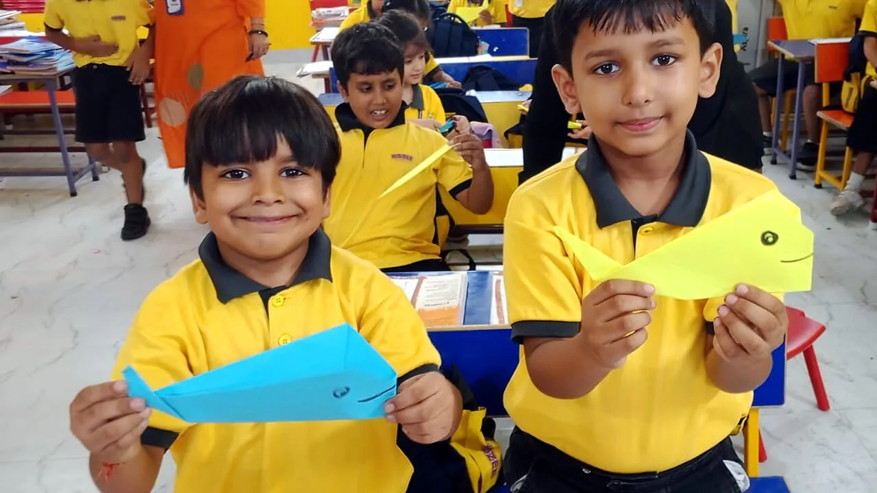 Two boys smiling as they hold up the paper fish they folded, one blue, one yellow, the class at its desks behind them