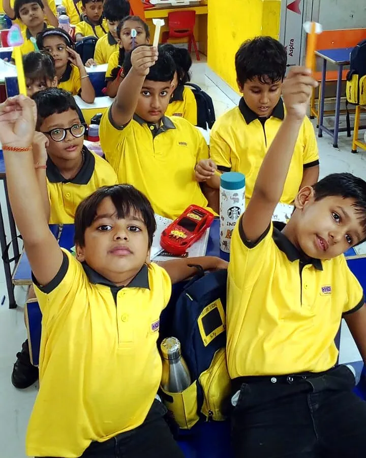 The class at its desks holding up the ice-cream sticks they made, a paper piece on each, two boys at the front with their arms high