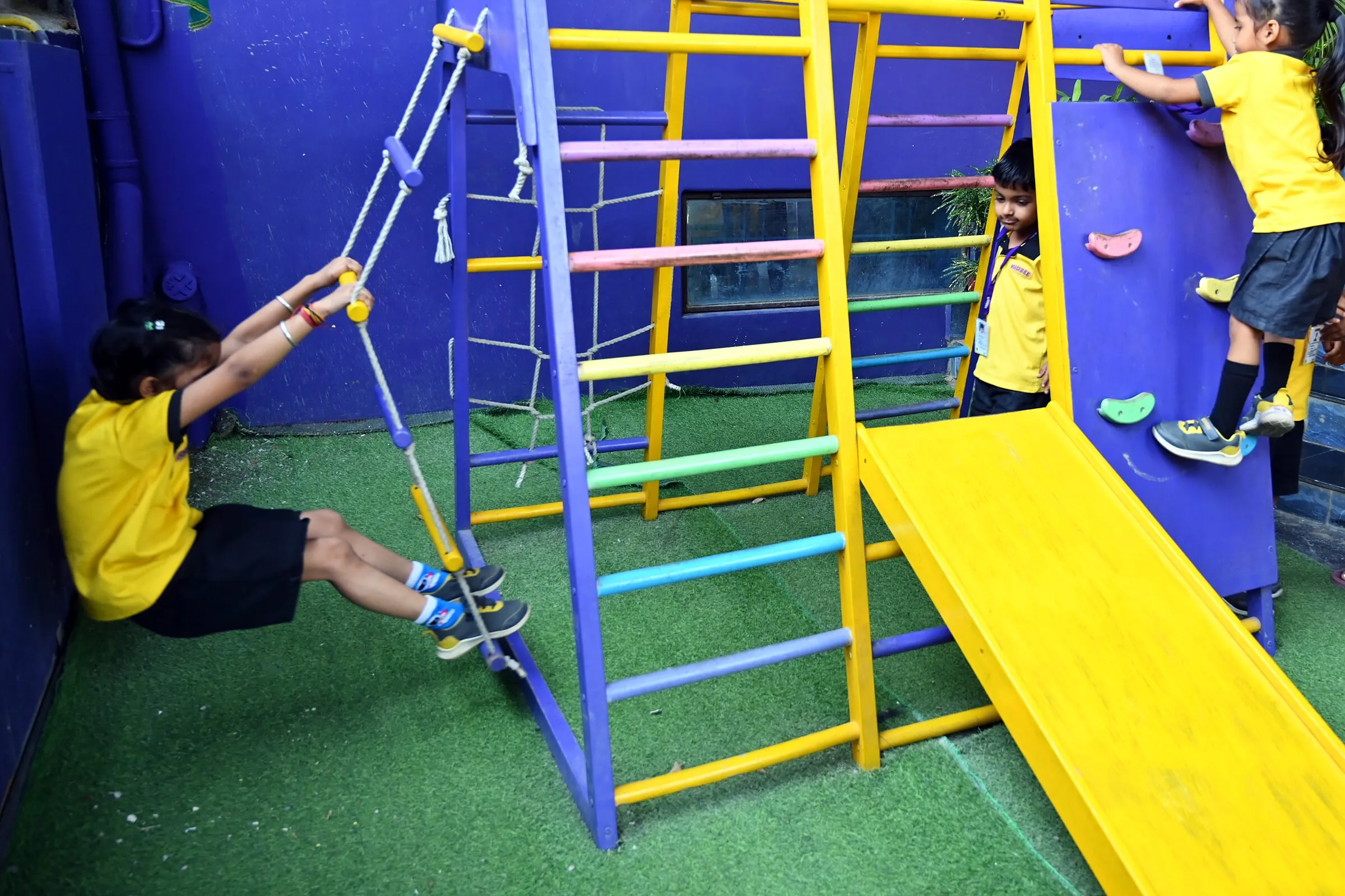 A girl climbing the rope ladder of the climbing frame, a girl at the wall beyond the slide