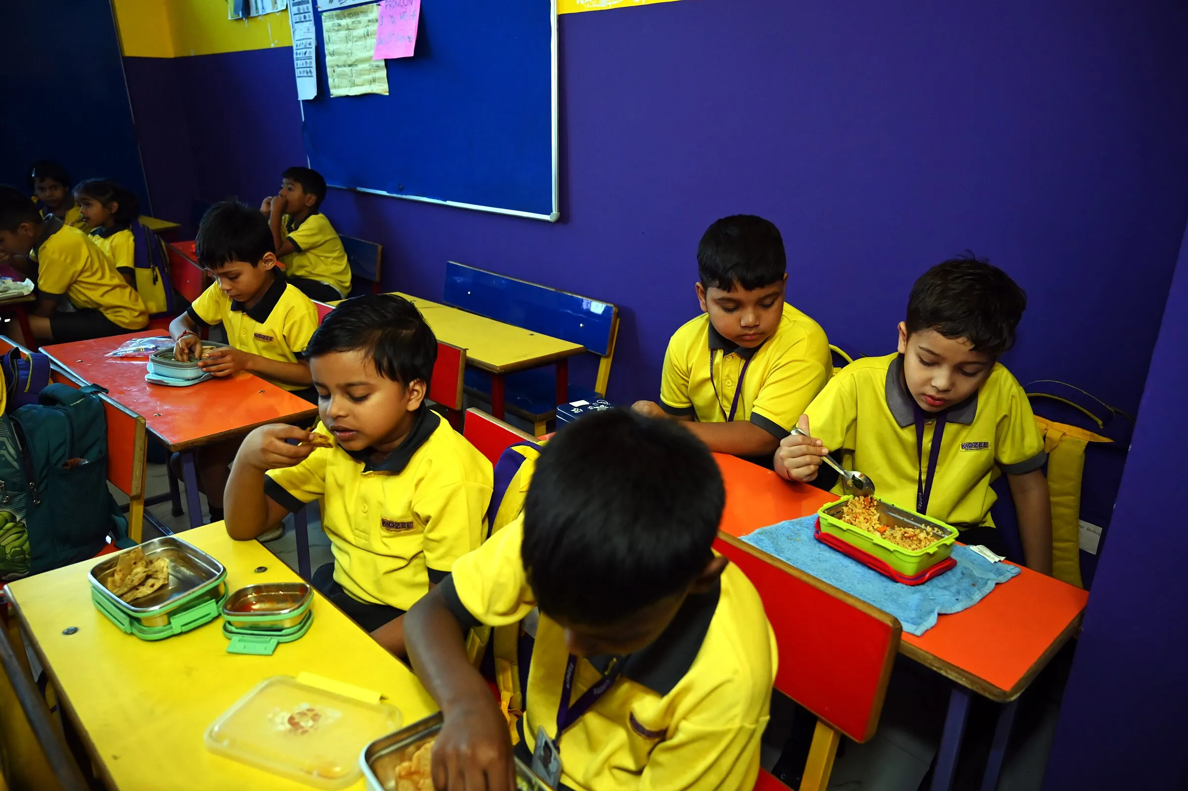 Children at their desks eating from their tiffin boxes, a boy looking up
