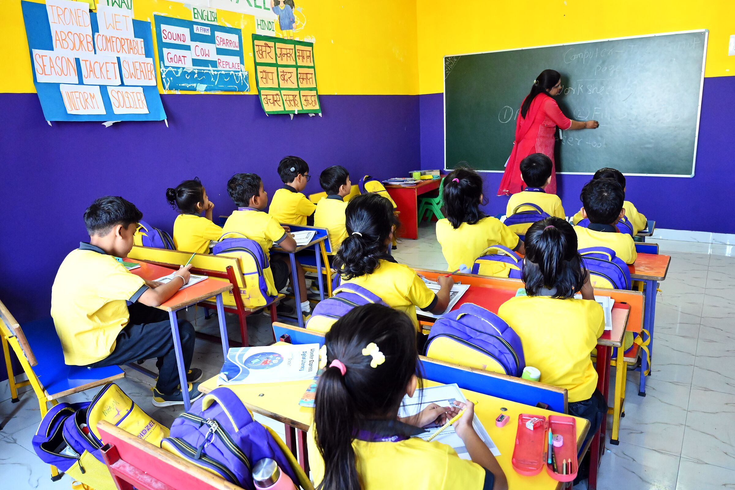 A class at work from the back of the room, children at their desks and the teacher writing on the board