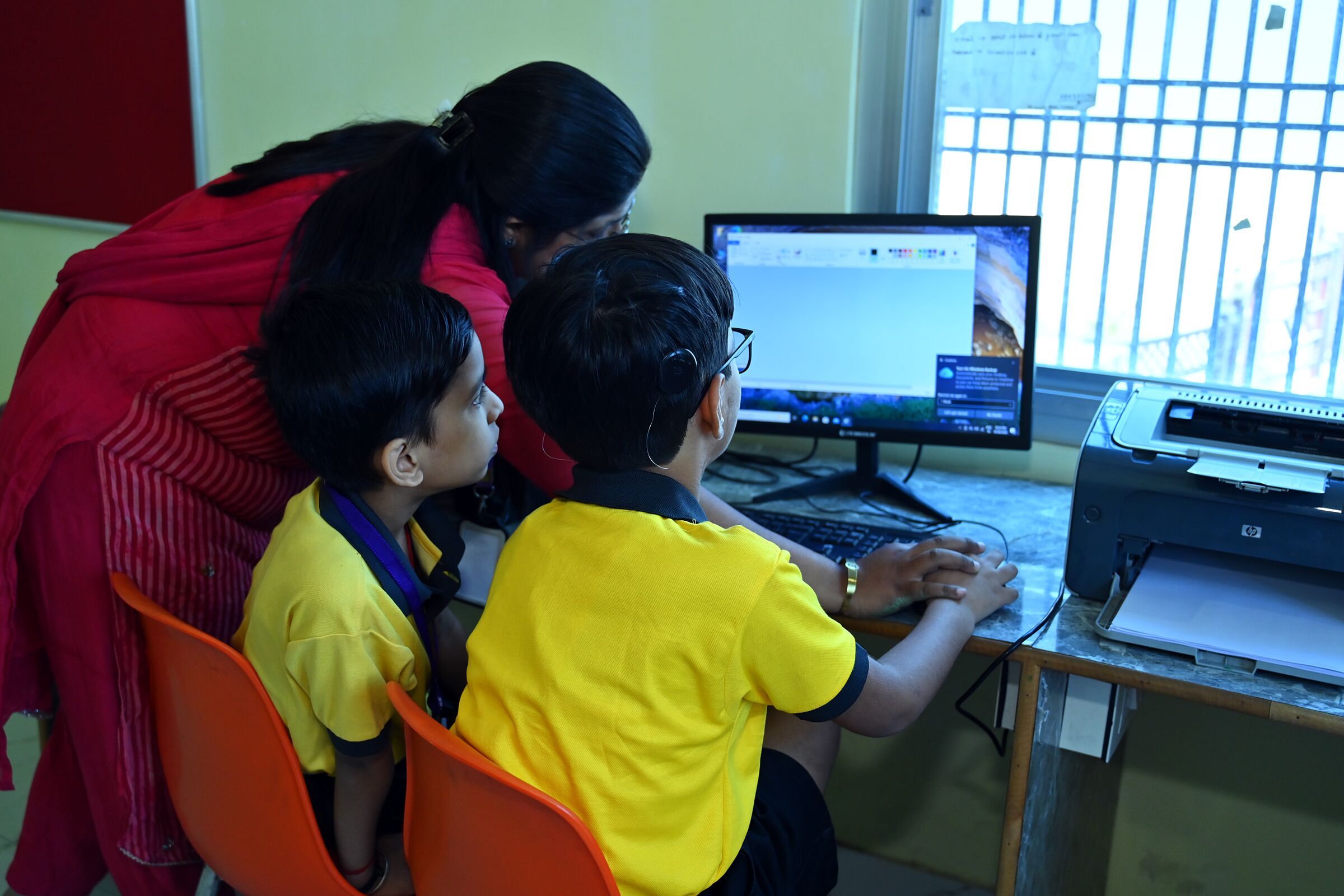 The computer lab: a teacher leaning in to guide two boys at a computer