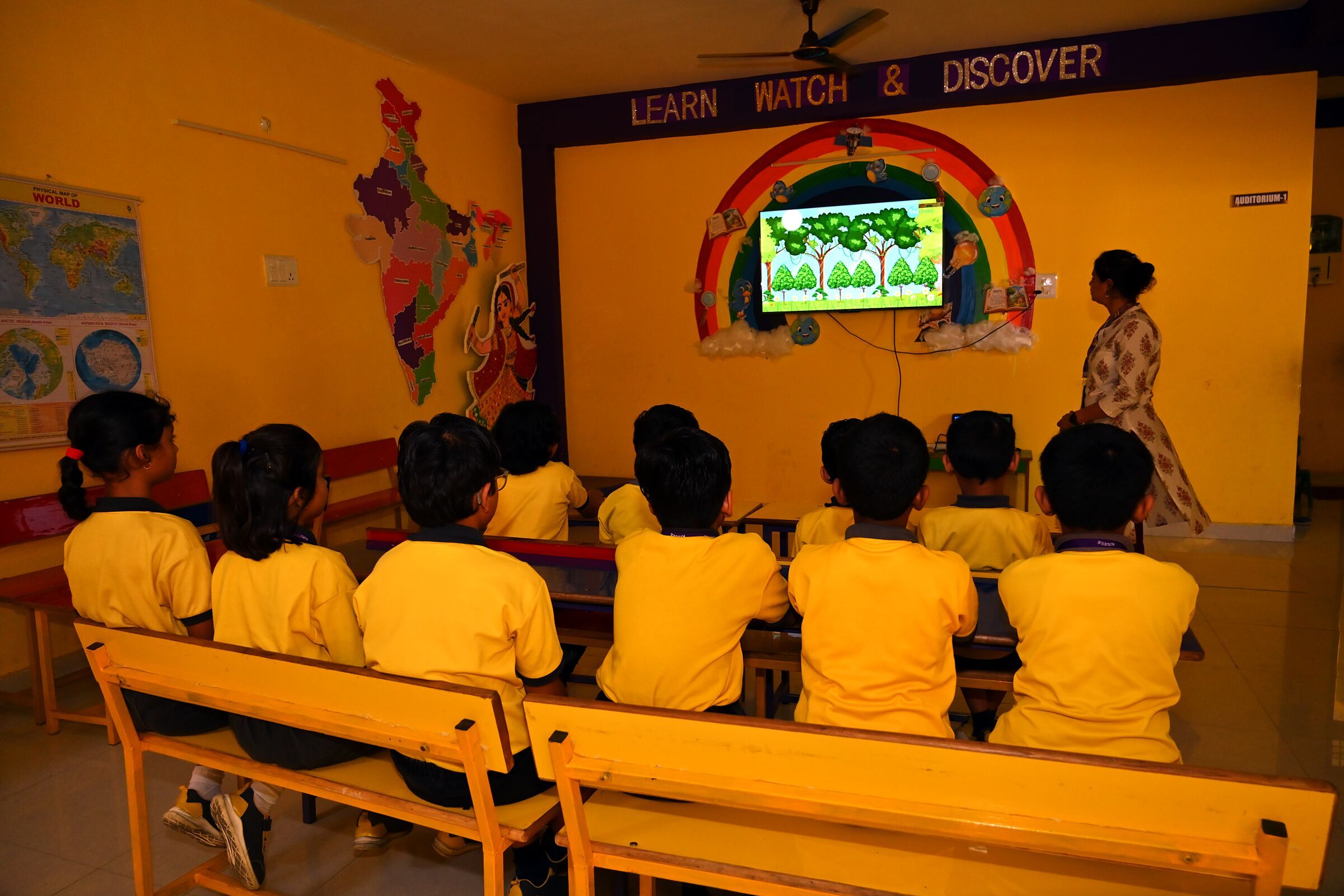 A class on its benches watching a learning programme on the smart TV, under the room's rainbow