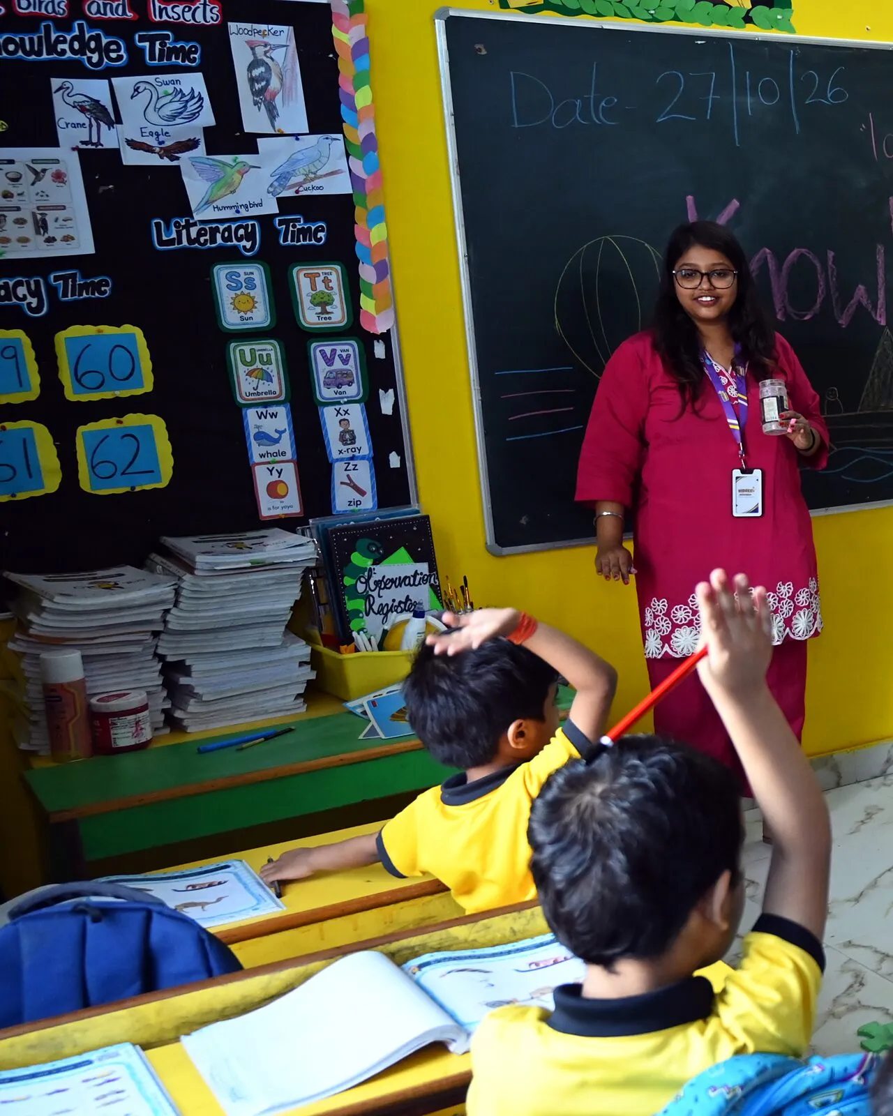 The number cards on the classroom wall beside the blackboard, a teacher standing by, a boy's hand up to answer