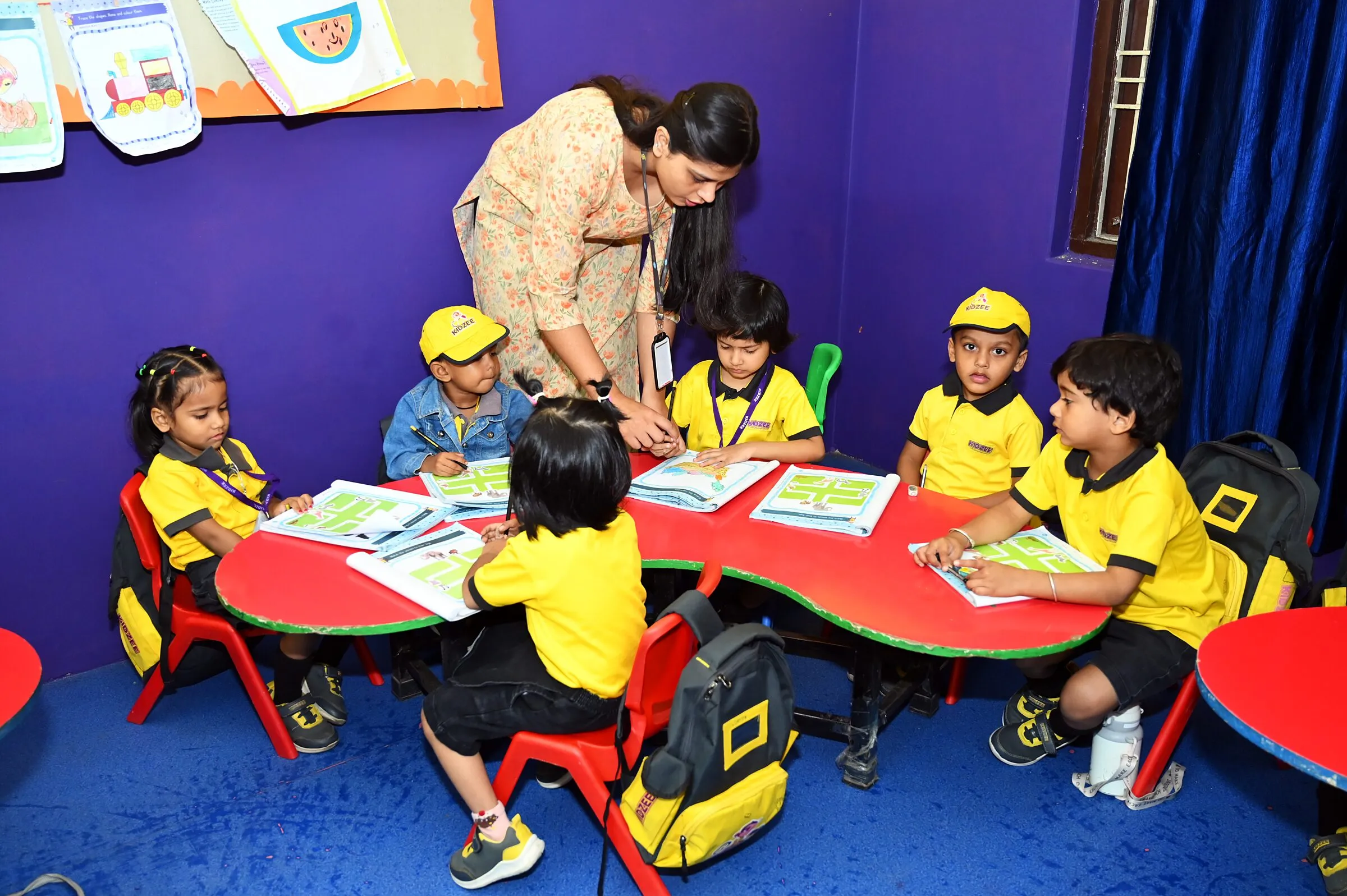 Children tracing the mazes in their workbooks round a red table, their teacher bending to help