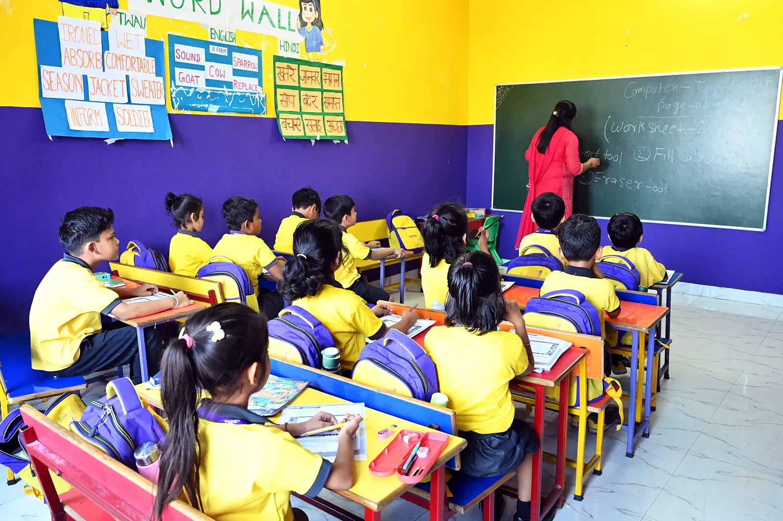 A class at its desks from the back, the teacher writing on the board beside the class's word wall