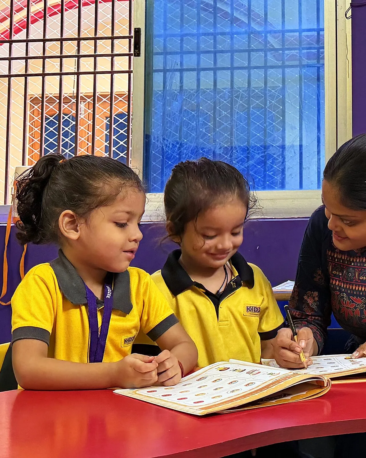 Two girls smiling over their workbook as one writes, their teacher beside them