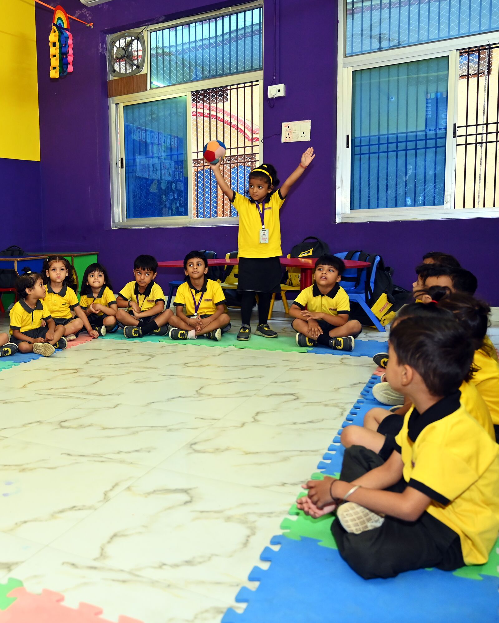 Circle time: a girl standing up with the ball, her arm high, the class sitting round the mats