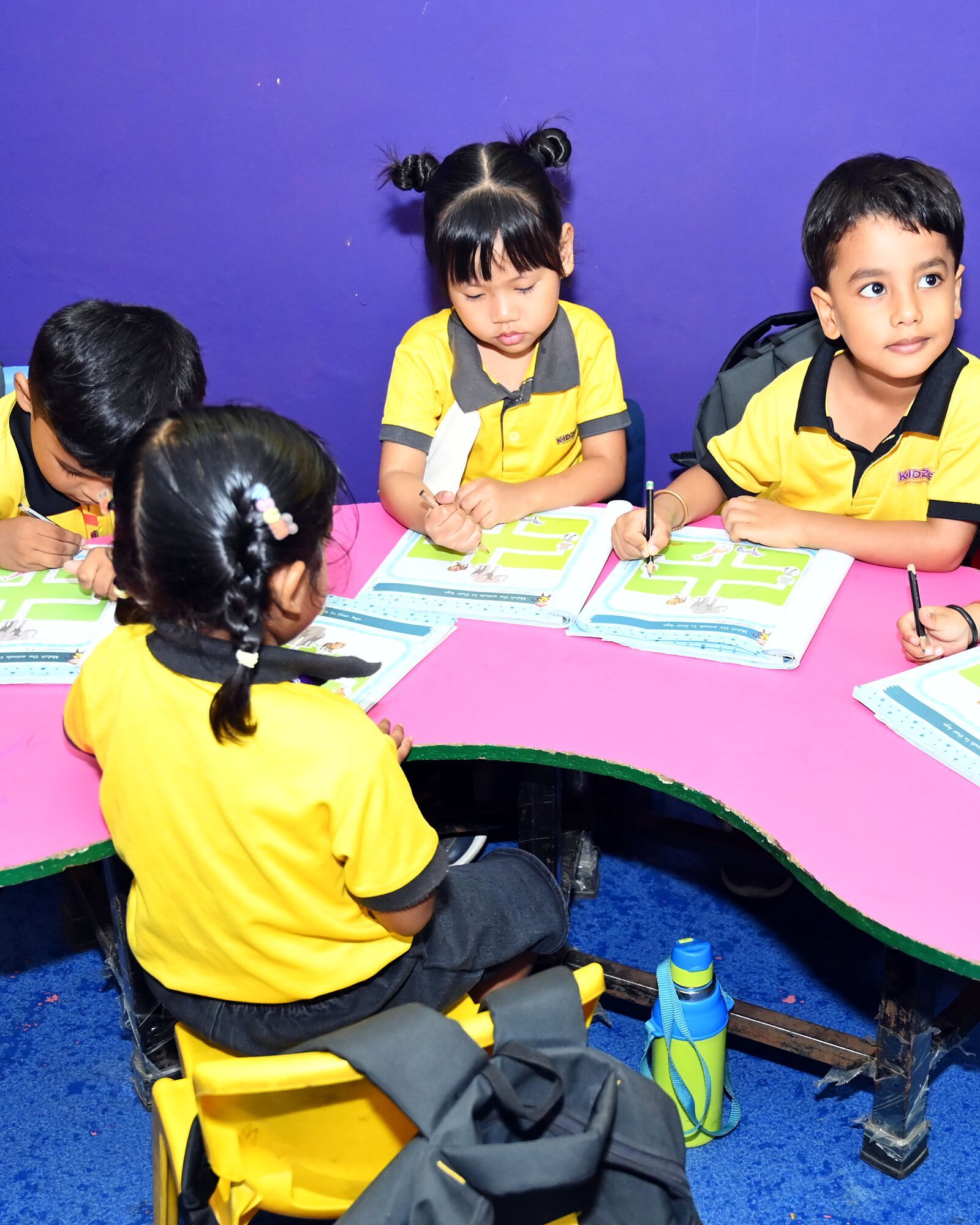 Children at work in their maze books round a pink table, seen over a girl's shoulder, a boy looking up