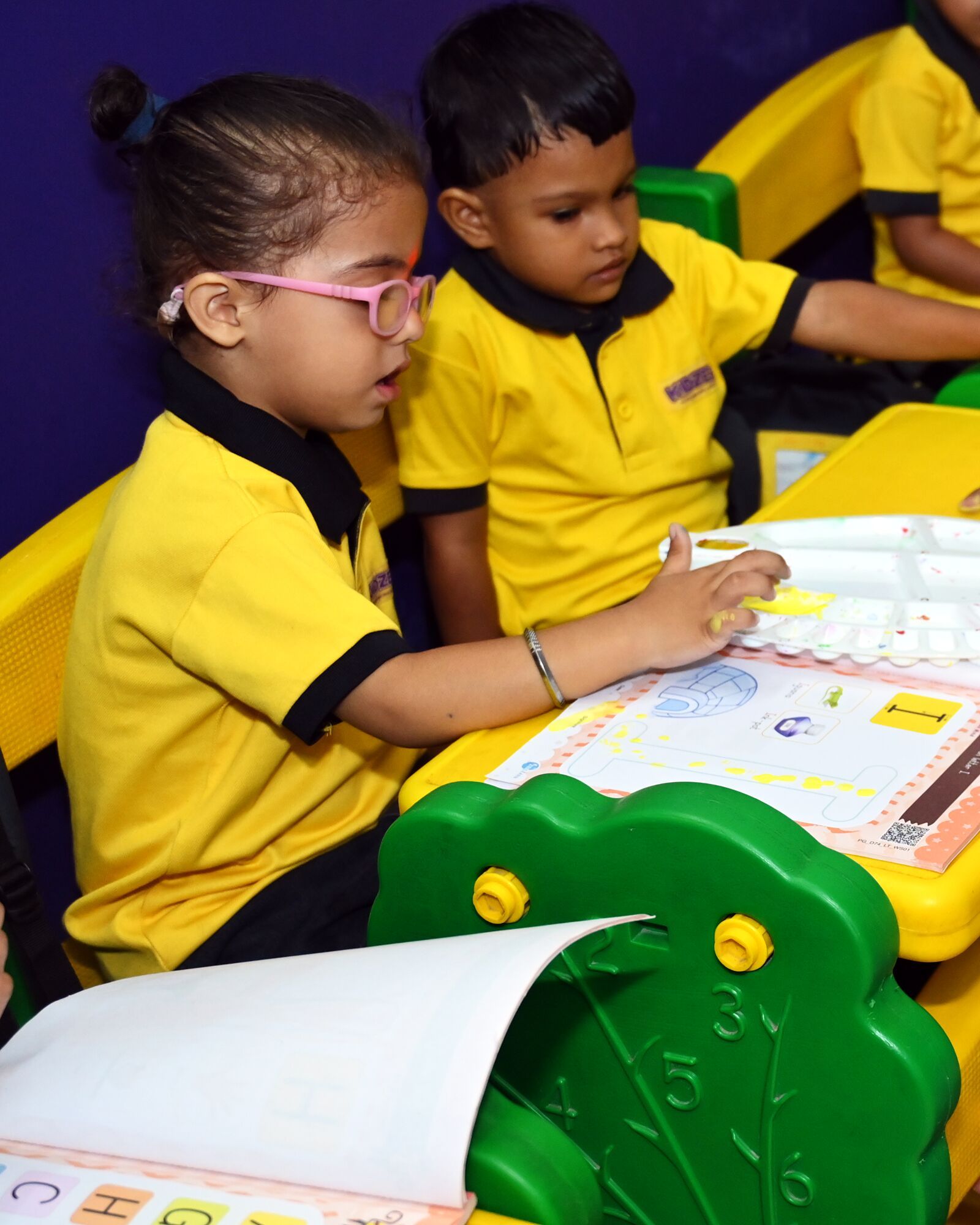 A girl in pink glasses dipping her fingers in the paints over her workbook, a boy beside her watching