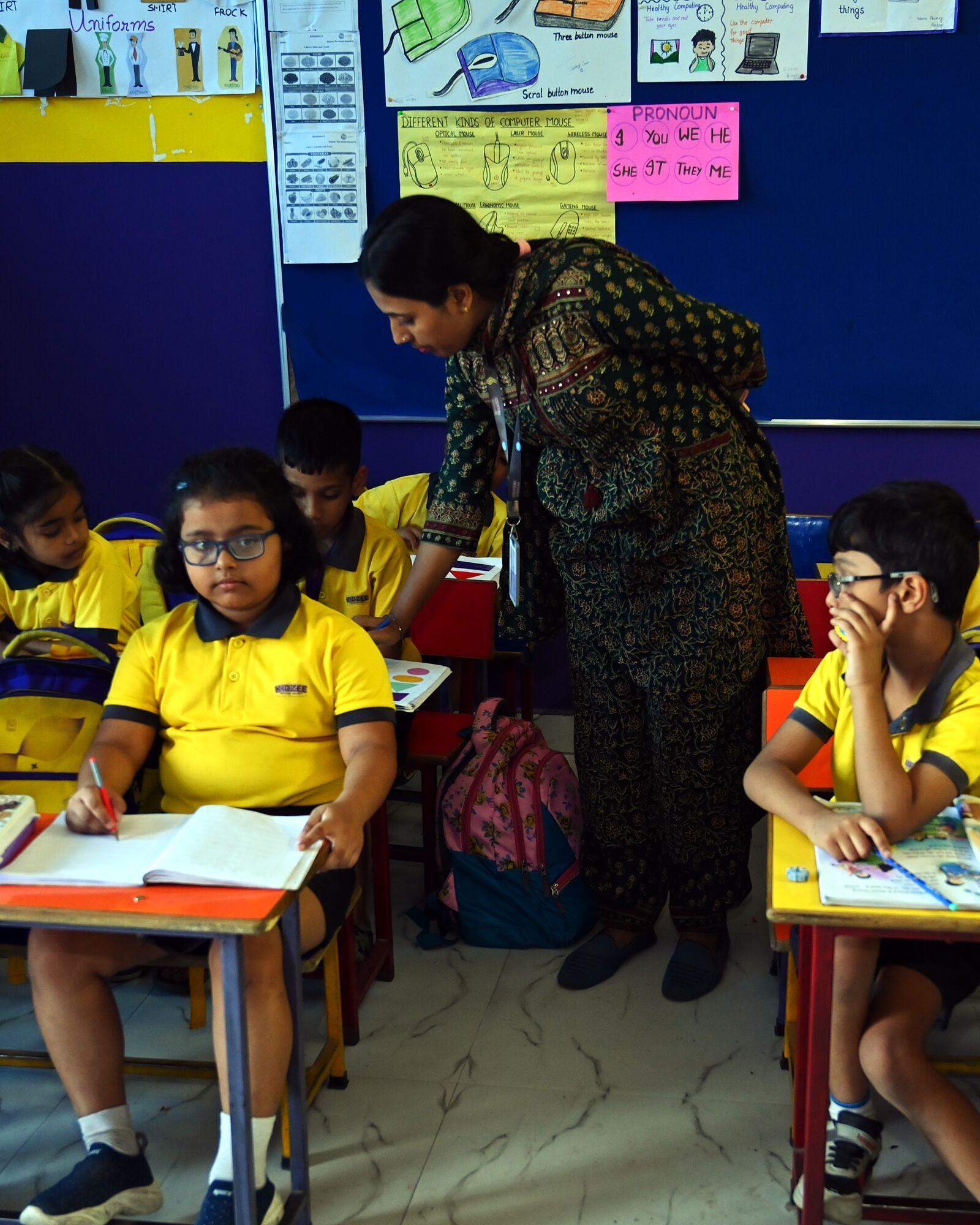 Children reading at their desks, their teacher bending between them to help