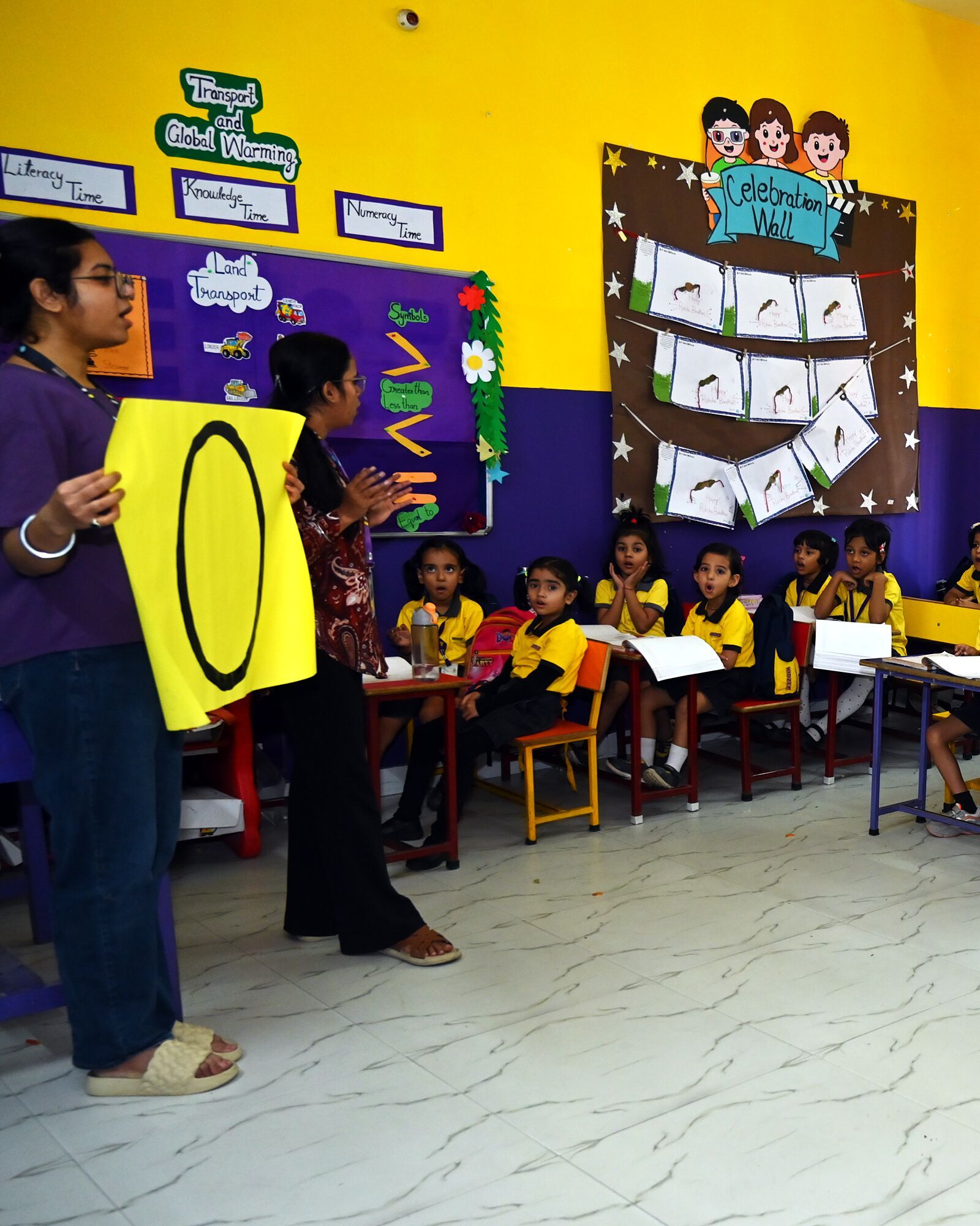 A teacher holding up a big zero to the class at its desks, a second teacher beside her